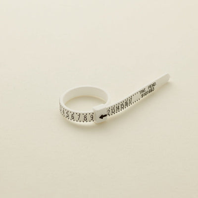 A white plastic zip-tie like ring sizer with black numbers, shown on a solid light beige surface.