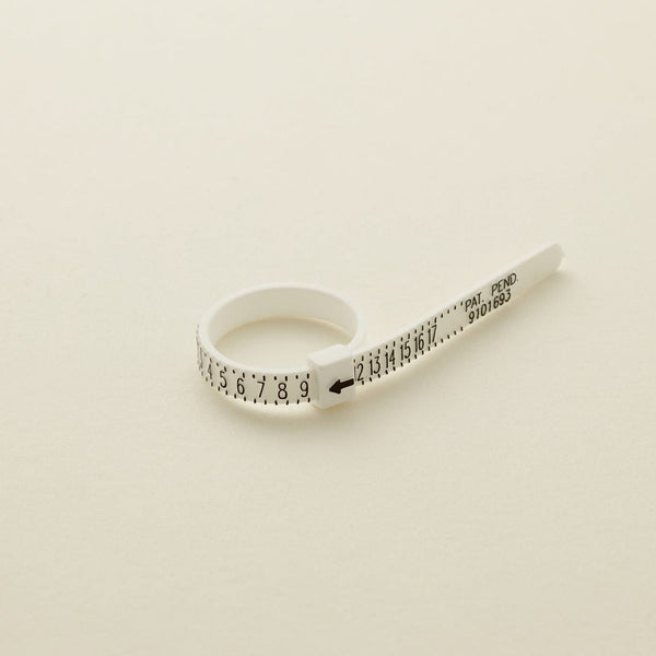 A white plastic zip-tie like ring sizer with black numbers, shown on a solid light beige surface.
