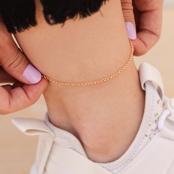 A close-up of a person wearing a gold round link chain anklet with white sneakers, with a solid beige background.