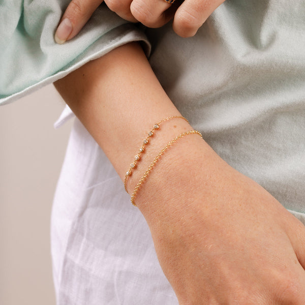 A close-up of a person's wrist wearing a gold round link chain bracelet and a gold delicate chain bracelet with seven round bezel set clear gemstones in a line.