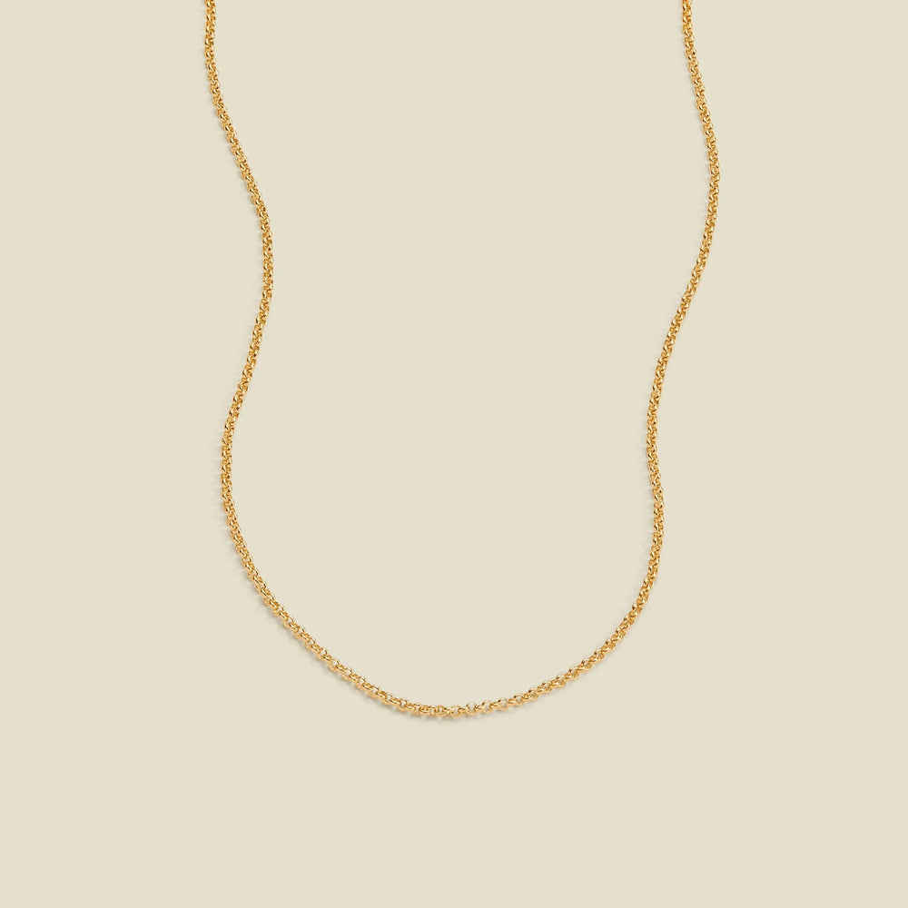 A delicate gold chain necklace with a round link pattern, displayed against a solid light beige background.