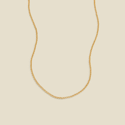 A delicate gold chain necklace with a round link pattern, displayed against a solid light beige background.