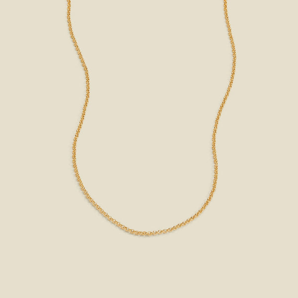 A delicate gold chain necklace with a round link pattern, displayed against a solid light beige background.