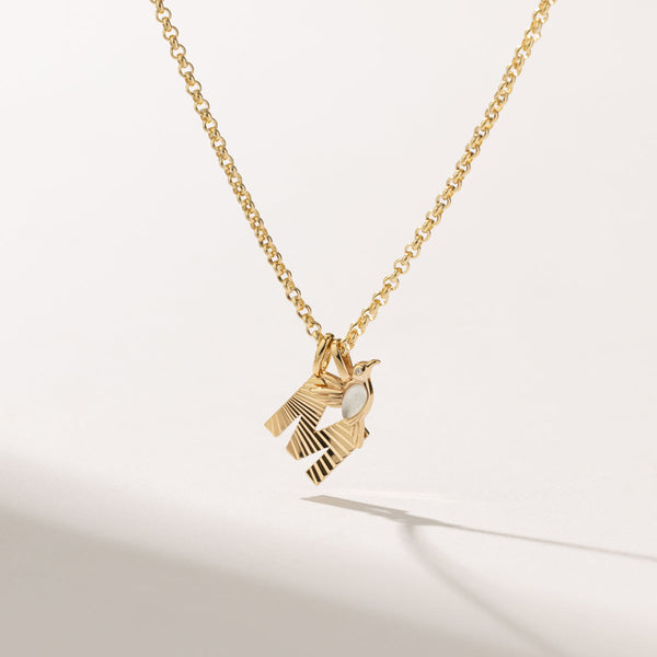 A round link gold chain necklace with a gold initial 'M' charm with a sunburst textured pattern, and a gold hummingbird charm with an opalescent gemstone on the bodice. set against a solid white background with shadows.