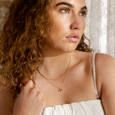 A close-up lifestyle image of a woman wearing a dainty round link gold chain necklace layered with a gold pendant chain necklace holding a gold dragonfly charm with natural gemstones on the wings.