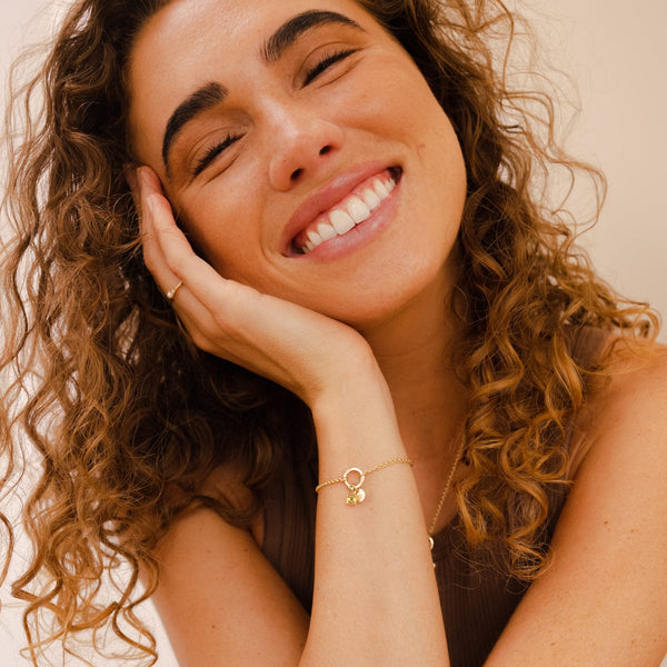 A woman resting her hand against her face, showcasing her wrist wearing a gold chain bracelet with a round ring in the center, holding a flat round disc charm and a square light-green gemstone charm.