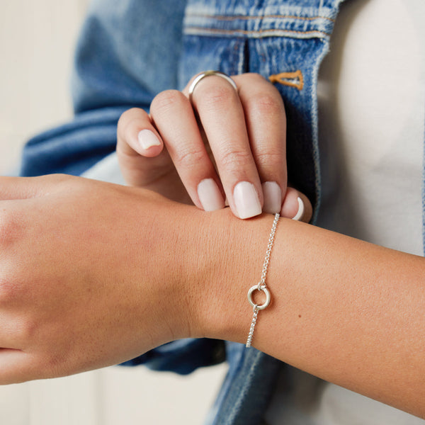 A person's wrist wearing a silver chain bracelet with a round ring in the center, paired with a white t-shirt layered with a dark blue jean jacket.