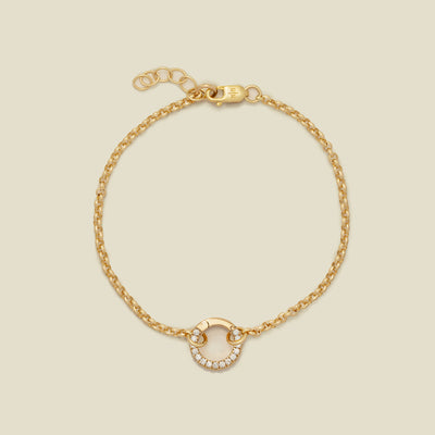 A gold circular link chain bracelet with a round ring in the center embellished with clear gemstones, shown against a solid light beige background.