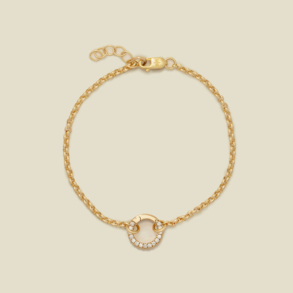A gold circular link chain bracelet with a round ring in the center embellished with clear gemstones, shown against a solid light beige background.