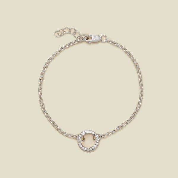 A silver circular link chain bracelet with a round ring in the center embellished with clear gemstones, shown against a solid light beige background.