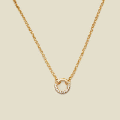 A gold circular link chain necklace with a round ring embellished with clear accent stones, shown on a solid light beige background.