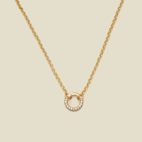 A gold circular link chain necklace with a round ring embellished with clear accent stones, shown on a solid light beige background.