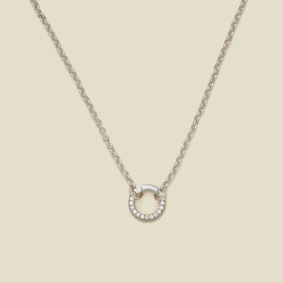 A silver circular link chain necklace with a round ring in the center embellished with clear accent stones, shown on a solid light beige background.
