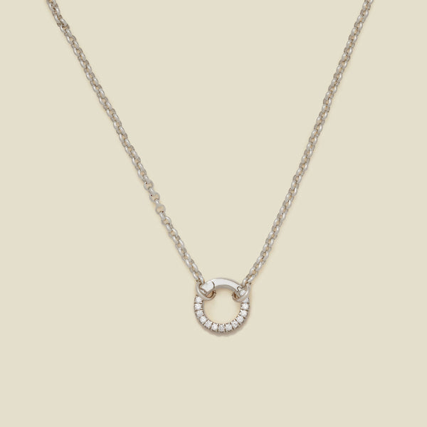A silver circular link chain necklace with a round ring in the center embellished with clear accent stones, shown on a solid light beige background.