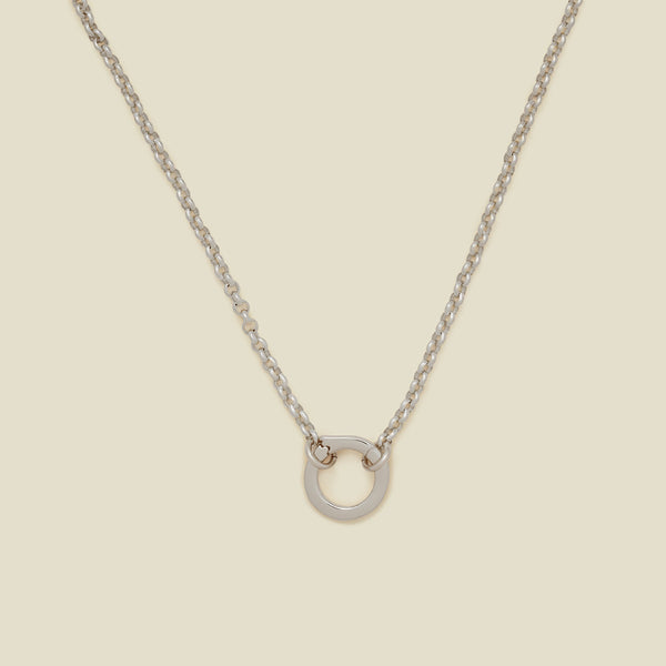A silver circular link chain charm necklace with a hinge-style round ring in the center, displayed on a solid beige background.