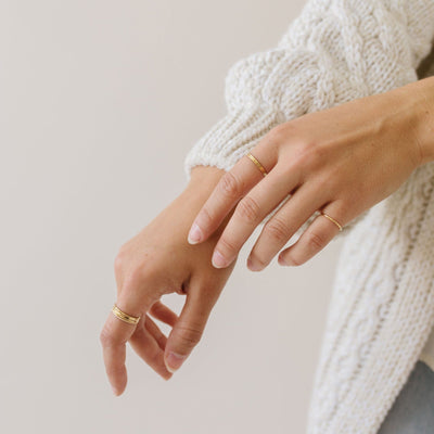 A person's hands held out in front of them, showcasing multiple thin gold stacked wire rings, shown on a solid off-white background.