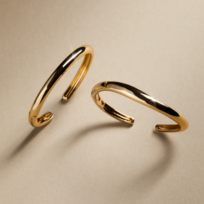 Two polished gold bangle bracelets, one with an organic curve, set against a textured beige background with moody lighting and shadows.