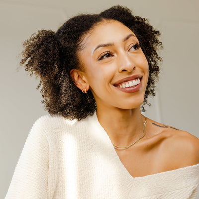 A person wearing two wide flat necklaces in gold and silver, paired with two gold hoop earrings and set against a light grey background.