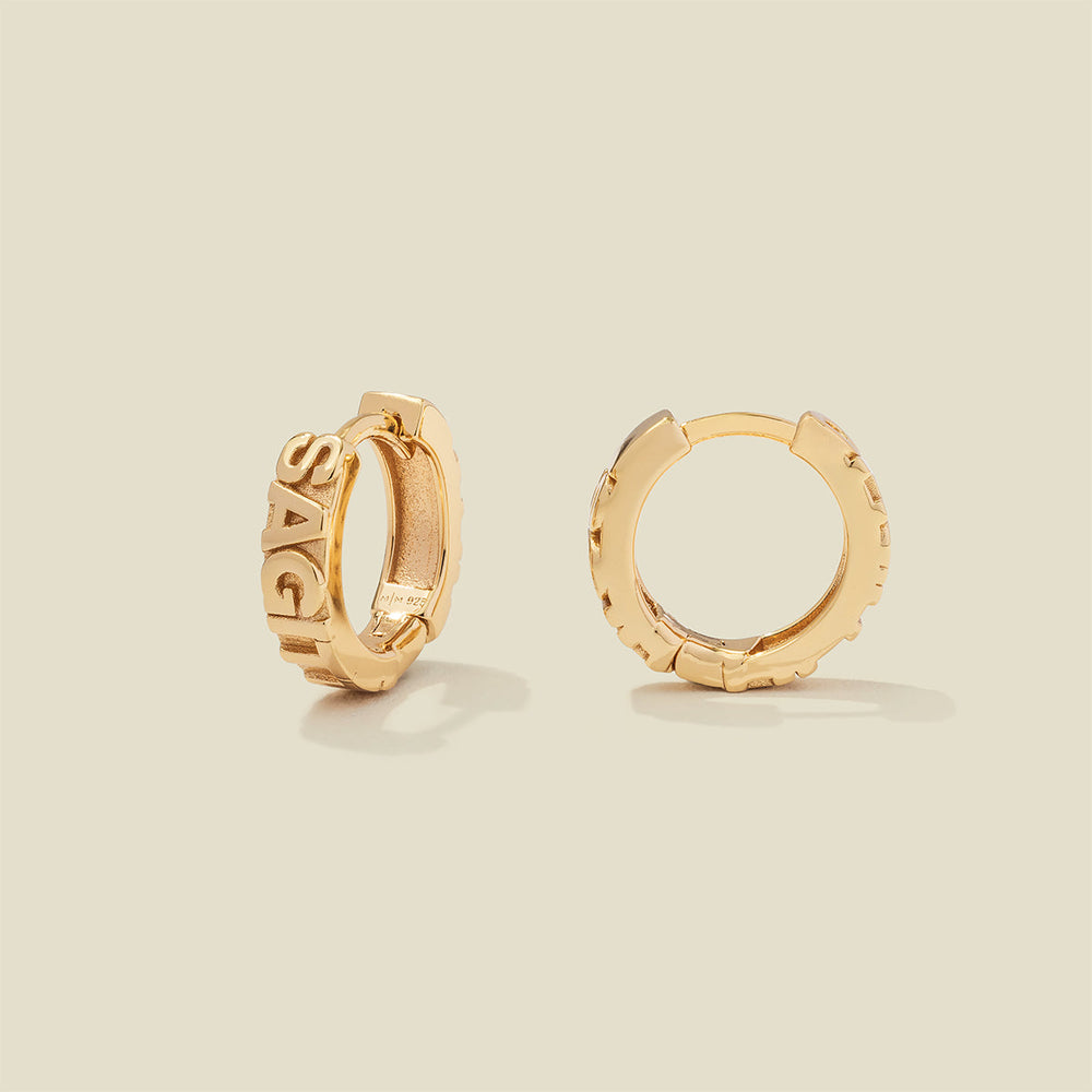 A pair of gold huggie hoop earrings on a light beige background. One earring is shown from the side, displaying a polished outer surface, while the other is shown from the front, showcasing the word 'SAGITTARIUS' in raised letters.