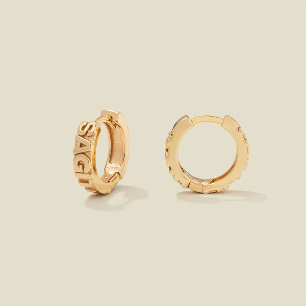 A pair of gold huggie hoop earrings on a light beige background. One earring is shown from the side, displaying a polished outer surface, while the other is shown from the front, showcasing the word 'SAGITTARIUS' in raised letters.