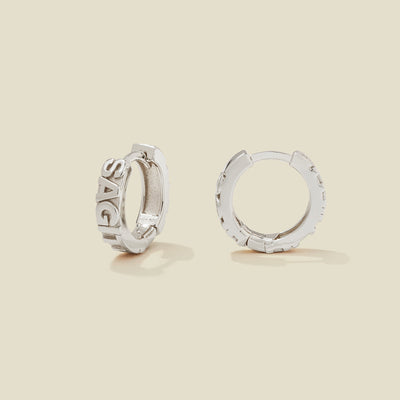 A pair of silver huggie hoop earrings on a light beige background. One earring is shown from the side, displaying a polished outer surface, while the other is shown from the front, showcasing the word 'SAGITTARIUS' in raised letters.