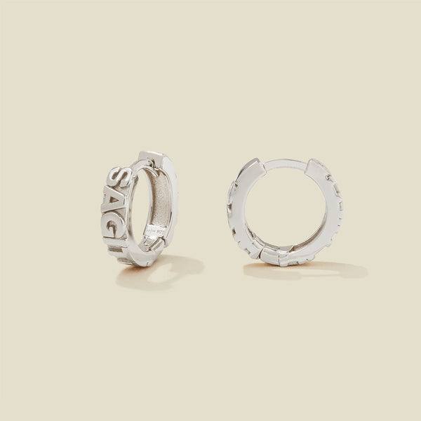 A pair of silver huggie hoop earrings on a light beige background. One earring is shown from the side, displaying a polished outer surface, while the other is shown from the front, showcasing the word 'SAGITTARIUS' in raised letters.