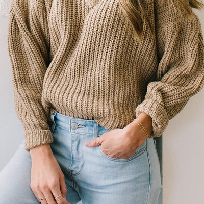 A person wearing light blue denim jeans and a light brown knit sweater, paired with two delicate gold chain bracelets with round polished beads.