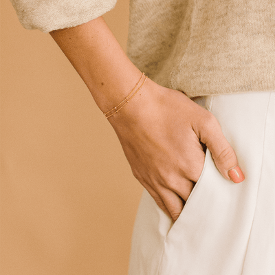 A hand resting in the pocket of white trouser pants, showcasing two delicate gold chain bracelets with round stationary gold beads.