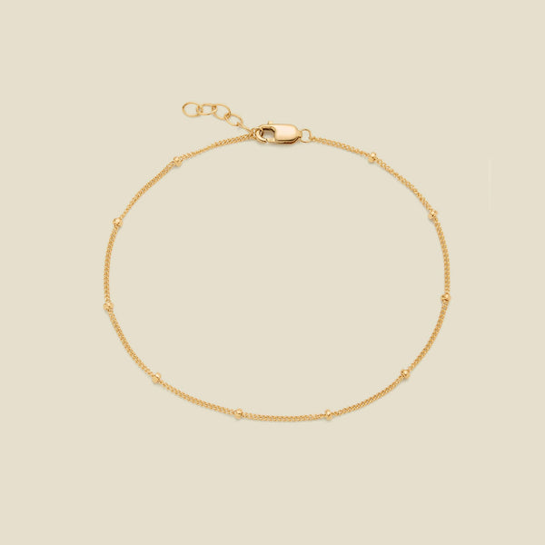 A delicate gold chain bracelet with small spaced beads and a lobster clasp, displayed on a solid light beige background.
