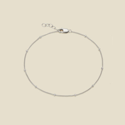 A delicate silver chain bracelet with small spaced beads and a lobster clasp, displayed on a solid light beige background.