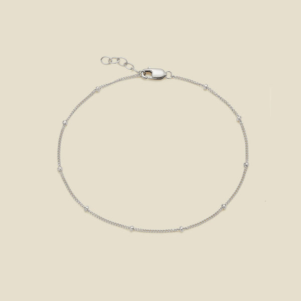A delicate silver chain bracelet with small spaced beads and a lobster clasp, displayed on a solid light beige background.