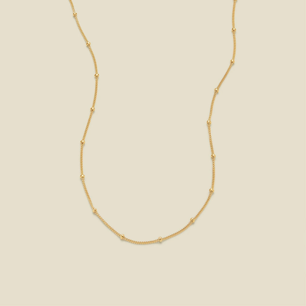 A delicate gold chain necklace with evenly spaced small round bead links, displayed on a solid light beige background.