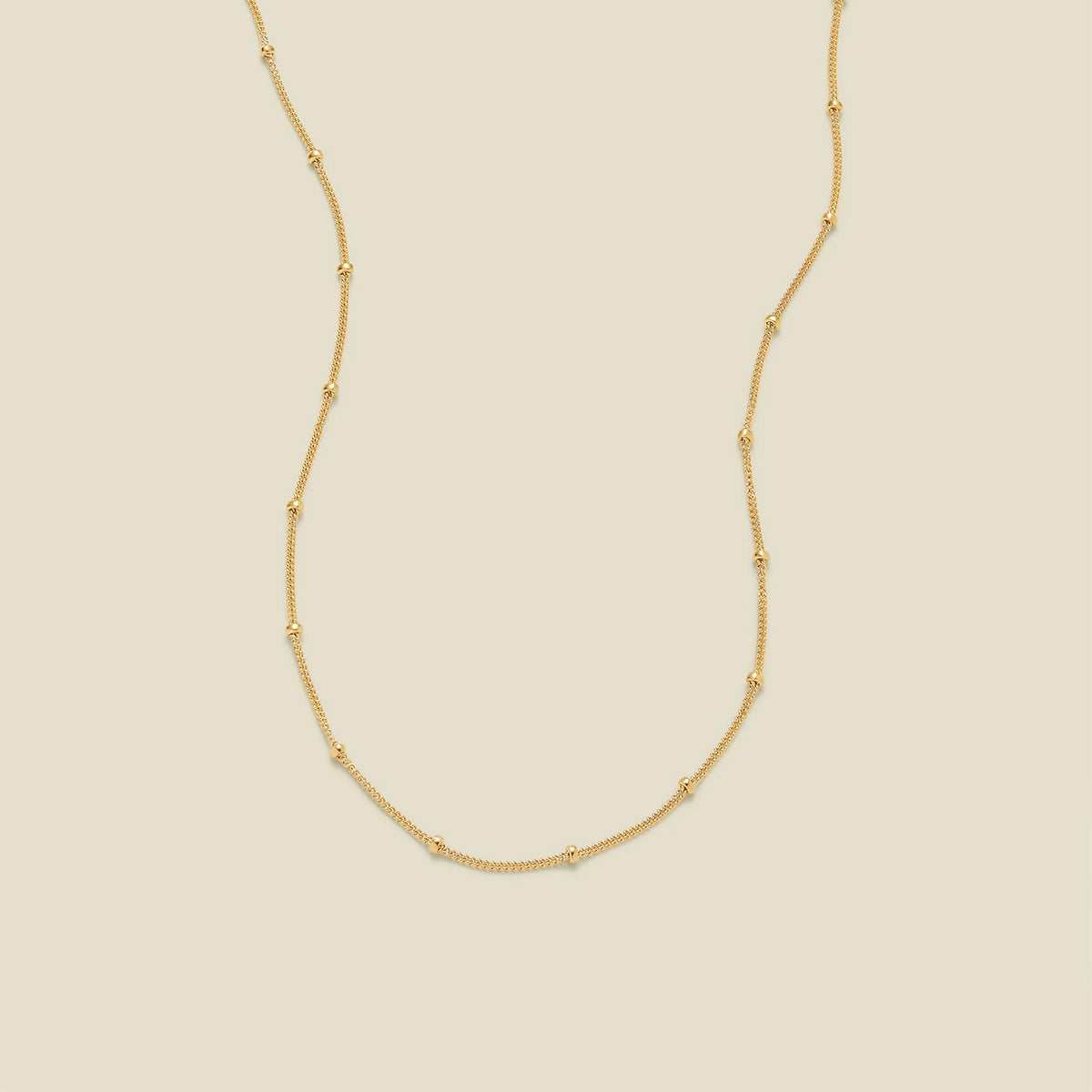 A delicate gold chain necklace with evenly spaced small round bead links, displayed on a solid light beige background.