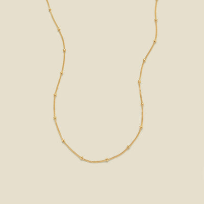 A delicate gold chain necklace with evenly spaced small round bead links, displayed on a solid light beige background.