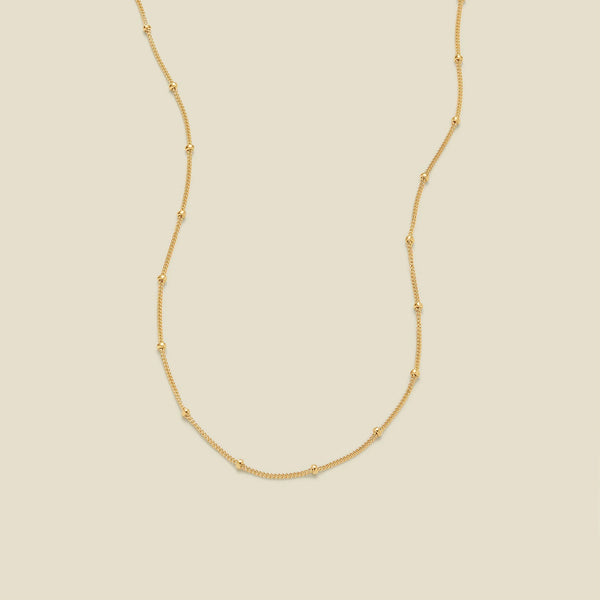 A delicate gold chain necklace with evenly spaced small round bead links, displayed on a solid light beige background.