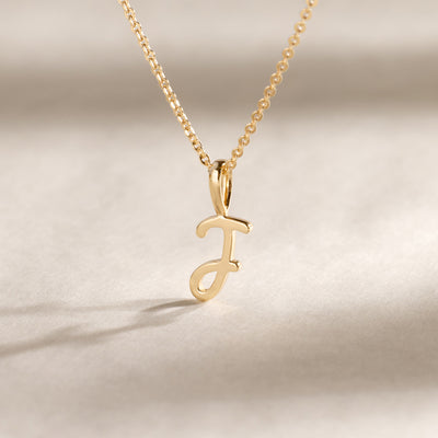 Gold chain with a gold J charm suspended against a plain beige background.