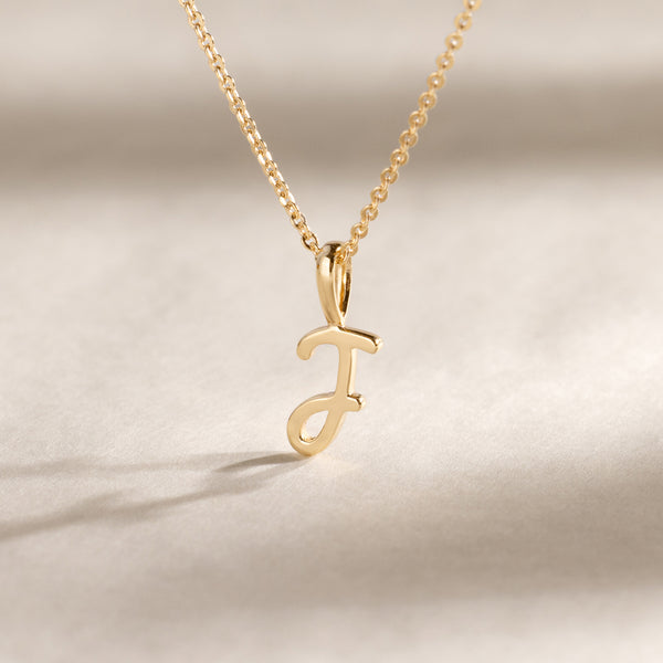 Gold chain with a gold J charm suspended against a plain beige background.