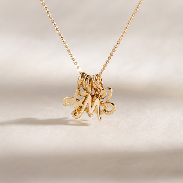Close-up of a gold chain necklace with multiple cursive initial charms clustered together, on a neutral background.