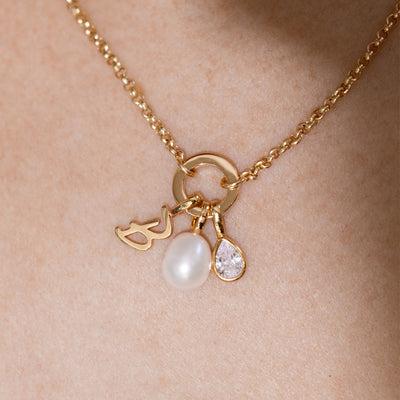 A close-up of a gold necklace worn on a person’s neck. It features a circular link lock holding three small charms: a cursive initial "B" charm, a smooth oval pearl, and a teardrop-shaped clear gemstone.