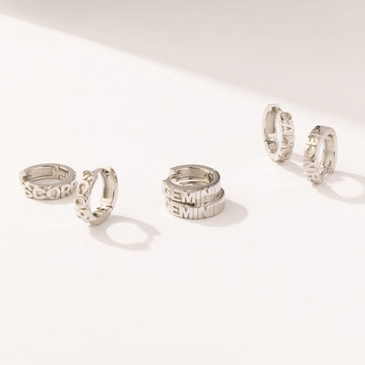 Three pairs of silver huggie hoops on a solid white surface, each with the name of a zodiac sign in raised lettering on the front.