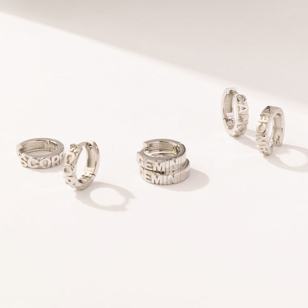 Three pairs of silver huggie hoops on a solid white surface, each with the name of a zodiac sign in raised lettering on the front.
