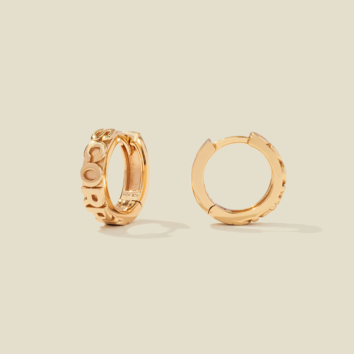 A pair of gold huggie hoop earrings on a light beige background. One earring is shown from the side, displaying a polished outer surface, while the other is shown from the front, showcasing the word 'SCORPIO' in raised letters.