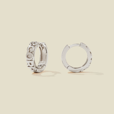 A pair of silver huggie hoop earrings on a light beige background. One earring is shown from the side, displaying a polished outer surface, while the other is shown from the front, showcasing the word 'SCORPIO' in raised letters.