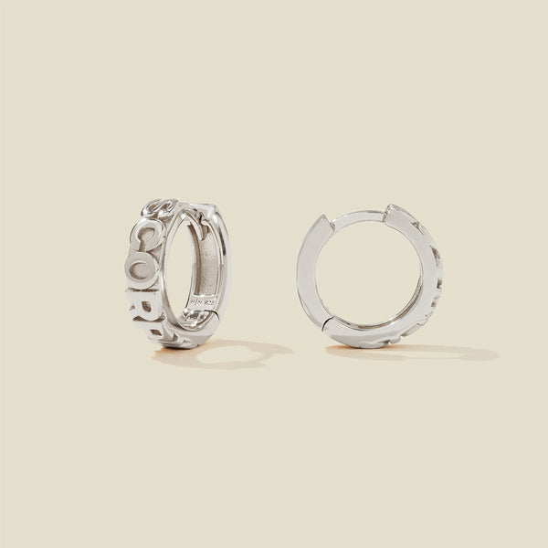 A pair of silver huggie hoop earrings on a light beige background. One earring is shown from the side, displaying a polished outer surface, while the other is shown from the front, showcasing the word 'SCORPIO' in raised letters.