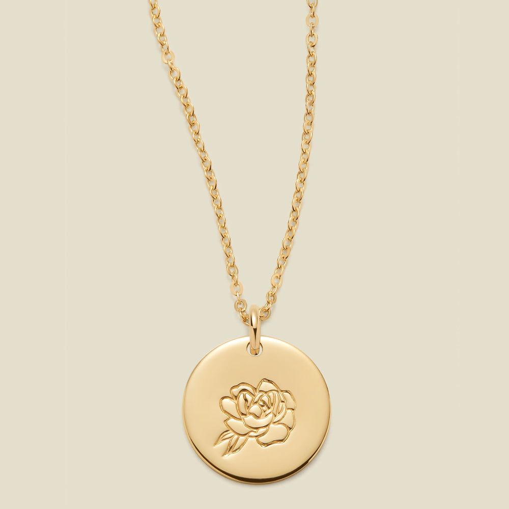A gold chain necklace with a round gold disc pendant engraved with a peony, shown against a solid light beige background.