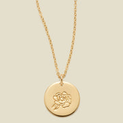 A gold chain necklace with a round gold disc pendant engraved with a peony, shown against a solid light beige background.