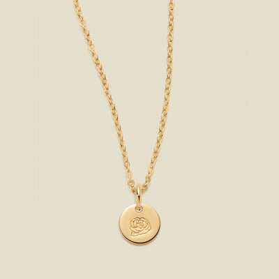 A gold chain necklace with a round gold disc pendant featuring an engraved peony flower design, displayed against a solid light beige background.