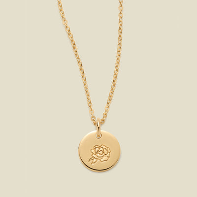 A gold chain necklace with a round gold disc pendant featuring an engraved peony flower design, displayed against a solid light beige background.