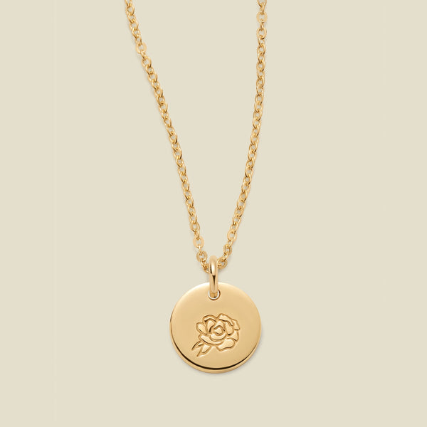 A gold chain necklace with a round gold disc pendant featuring an engraved peony flower design, displayed against a solid light beige background.
