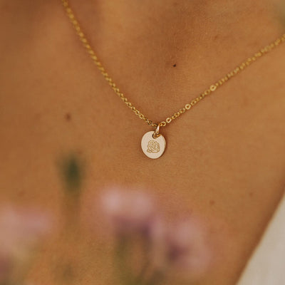 A close-up of a person wearing a gold pendant chain necklace with a round flat disc charm, engraved with a peony flower with pink flowers blurred in the foreground.